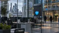 des bureaux de Barclays à Londres, avec des postes de travail inoccupés, des écrans éteints et deux professionnels marchant vers un hall vitré. Les enseignes bleues de la banque et les immeubles du quartier financier apparaissent à l’arrière-plan. Le visuel accompagne un article sur les suppressions de postes et la transformation technologique du groupe bancaire.
