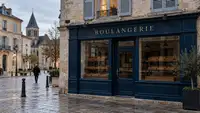 Façade bleu nuit d’une boulangerie fermée aux étagères et paniers à pain vides, dans un bourg français aux bâtiments en pierre, avec un passant sur la place pavée et un clocher en arrière-plan. Image générée par intelligence artificielle pour illustrer l’enquête Entreprisma sur l’évolution des boulangeries en France entre 2015 et 2025, leur présence dans les petites communes et les enjeux de maintien des commerces de proximité.