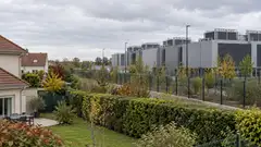 Illustration d’un centre de données contemporain avec des équipements de refroidissement, situé au-delà d’une clôture et à proximité de maisons avec jardins. Cette scène générique évoque les enjeux de voisinage du projet de Ballainvilliers, sans représenter son architecture ni son implantation exacte.