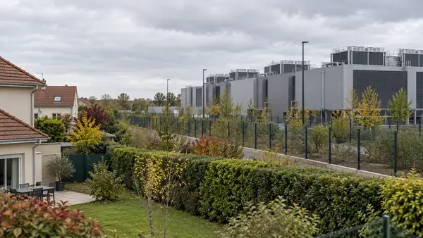 Illustration d’un centre de données contemporain avec des équipements de refroidissement, situé au-delà d’une clôture et à proximité de maisons avec jardins. Cette scène générique évoque les enjeux de voisinage du projet de Ballainvilliers, sans représenter son architecture ni son implantation exacte.