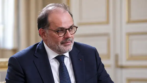 Portrait d’Emmanuel Moulin, gouverneur de la Banque de France, portant des lunettes rondes, une barbe grise, un costume bleu marine et une cravate sombre. Il apparaît de trois quarts dans un intérieur institutionnel clair à l’arrière-plan flouté. Illustration générée par intelligence artificielle pour un article sur ses déclarations concernant le budget 2027 et les critiques politiques qu’elles suscitent.