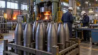 Un atelier de forge, avec des corps métalliques d’obus en cours de fabrication rangés au premier plan et deux ouvriers équipés de protections près d’une presse industrielle.