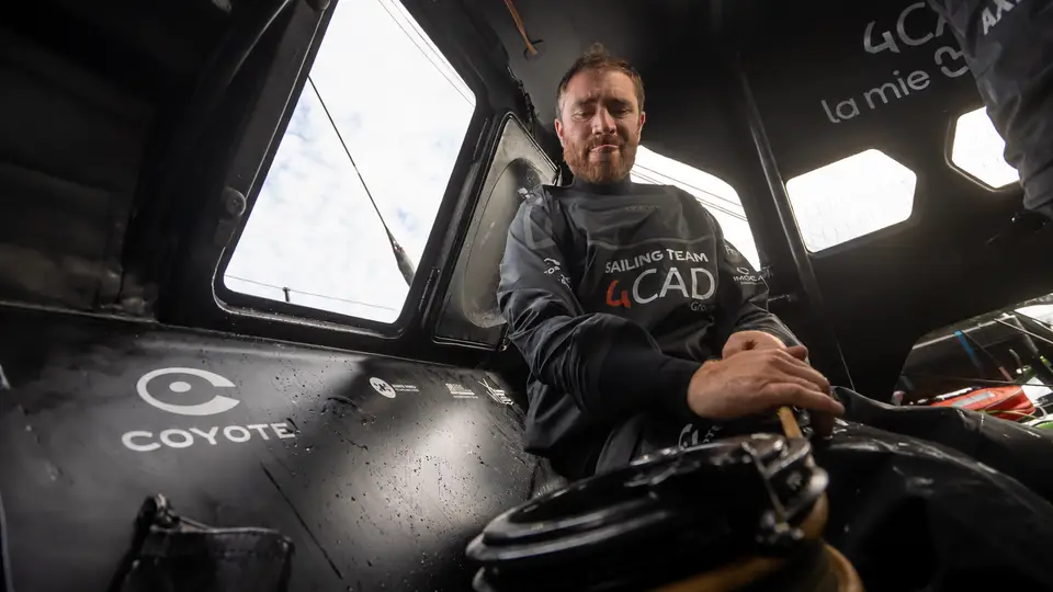 Benjamin Dutreux dans le cockpit d’un IMOCA aux couleurs de 4CAD Group, les mains posées sur un cordage près d’un winch. La photographie montre les équipements de manœuvre, les parois en carbone et les ouvertures du cockpit, au cœur de l’environnement de travail à bord d’un voilier de course au large.