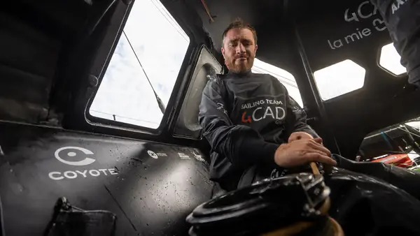 Benjamin Dutreux dans le cockpit d’un IMOCA aux couleurs de 4CAD Group, les mains posées sur un cordage près d’un winch. La photographie montre les équipements de manœuvre, les parois en carbone et les ouvertures du cockpit, au cœur de l’environnement de travail à bord d’un voilier de course au large.