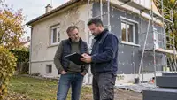 Un propriétaire et un artisan en tenue de chantier examinent un dossier devant une maison individuelle en cours de rénovation énergétique. La façade présente une partie ancienne et une partie recouverte de panneaux isolants, avec un échafaudage et des matériaux au premier plan. Illustration des travaux d’isolation extérieure et de leur préparation financière.