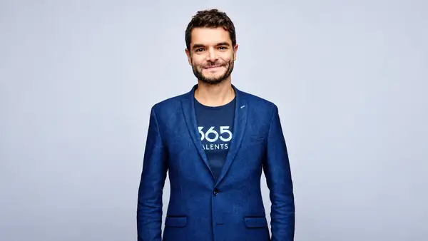 Portrait de Loïc Michel, cofondateur et directeur général de 365Talents, portant une veste bleue et un tee-shirt au logo de l’entreprise, sur fond gris clair.