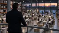 Vue d’un entrepôt de commerce en ligne au crépuscule : un responsable observe depuis une mezzanine les postes de préparation, les vêtements et les colis sur un convoyeur. Illustration de l’activité logistique évoquée dans l’article sur les résultats semestriels 2026 et la trésorerie de Showroomprivé.