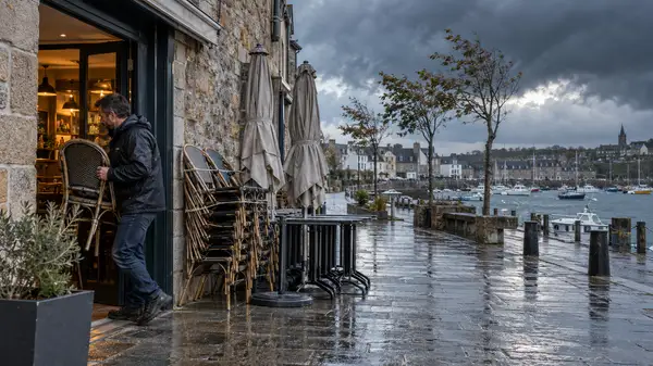 Un commerçant rentre une chaise dans un café donnant sur un port, sous un ciel sombre annonçant une tempête. Des chaises empilées, des tables regroupées et des parasols fermés illustrent la protection du mobilier de terrasse avant les vents forts.