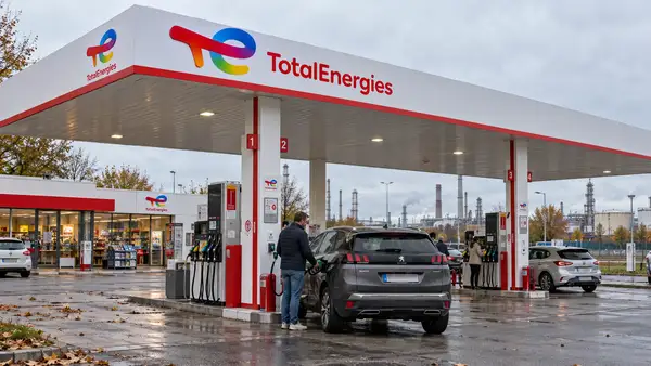 Illustration d’une station-service TotalEnergies avec son enseigne multicolore, plusieurs pompes et un automobiliste faisant le plein, tandis que des installations industrielles apparaissent à l’arrière-plan, pour illustrer les résultats du groupe et les différentes composantes du prix des carburants.