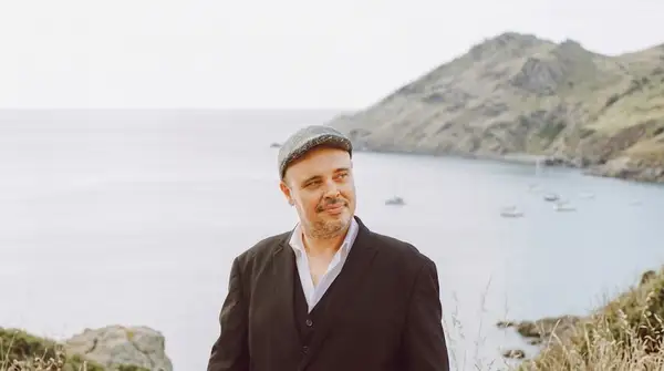Portrait professionnel d'Aurélien Gillet, fondateur de Mana, en costume noir et casquette tweed, regardant sur le côté devant une baie côtière et une colline verdoyante