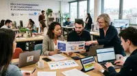 Une photographie réaliste de style reportage montre un groupe d'entrepreneurs diversifiés réunis autour d'une table en bois dans un bureau ouvert. Une femme d'âge mûr avec des lunettes sourit en montrant du doigt une brochure intitulée « GUIDE DE L'ENTREPRENEUR - Aides à la création d'entreprise » tenue par une jeune femme. Sur l'écran d'une tablette à côté d'elles, on lit « SUBVENTIONS RÉGIONALES ». Plusieurs ordinateurs portables, dont un affichant un site Web de la Bpifrance, sont ouverts. En arrière-plan, d'autres personnes travaillent dans l'espace de coworking, et un grand panneau mural porte l'inscription « PÔLE D'ACCOMPAGNEMENT ET DE FINANCEMENT À LA CRÉATION D'ENTREPRISE » avec les logos de France Travail, Bpifrance et Région Auvergne-Rhône-Alpes. L'image est en format 16:9 avec une lumière naturelle.