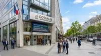 Un bâtiment tertiaire moderne à Paris affichant le logo d'une société de gestion immobilière (SCPI) avec un écran LED indiquant des chiffres clés du patrimoine. Devant l'immeuble, un groupe de professionnels en tenue d'affaires discute sur un trottoir animé.