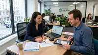 Un entrepreneur et une conseillère financière ou comptable discutent dans un bureau moderne et lumineux. Sur le bureau en bois sont posés des documents administratifs français, une tablette, une tasse de café et un ordinateur portable affichant une présentation intitulée « Comprendre le versement libératoire ». En arrière-plan, d'autres collaborateurs travaillent dans un espace ouvert avec de grandes baies vitrées donnant sur la ville.